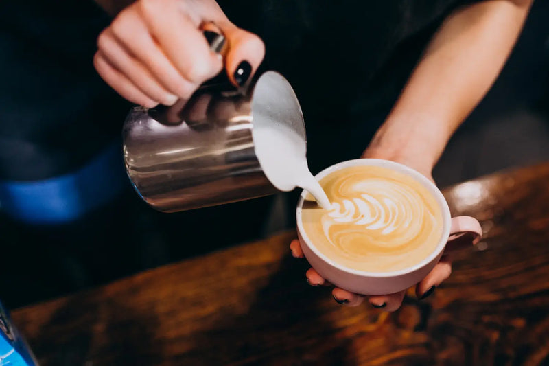 A person pours frothy milk into a latte, creating intricate latte art in a ceramic cup. The scene conveys warmth and craftsmanship, set on a wooden table.